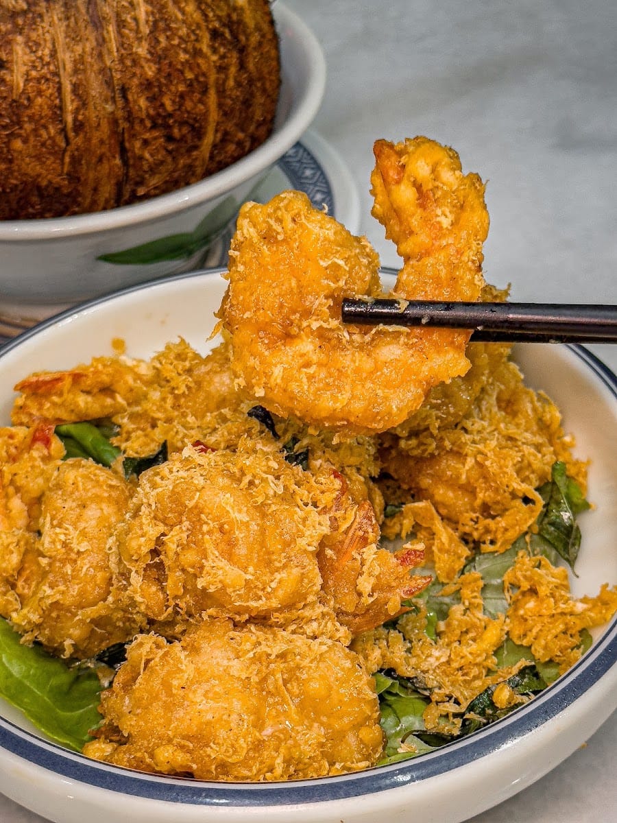 Fried prawns coated in golden cereal, lifted with chopsticks, with a coconut-shell bowl behind